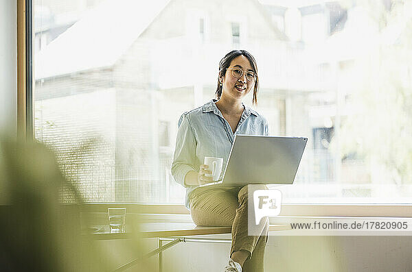 Smiling businesswoman with laptop and coffee cup sitting on desk