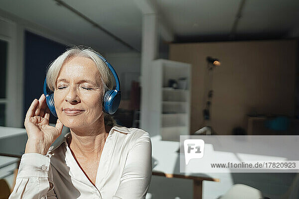Smiling senior woman with eyes closed listening music through wireless headphones in office