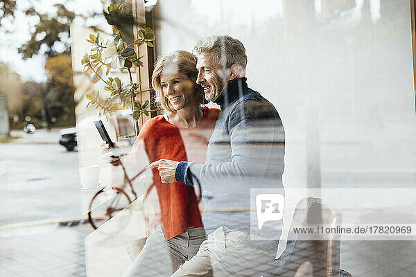 Smiling woman showing smart phone to man in cafe seen through glass
