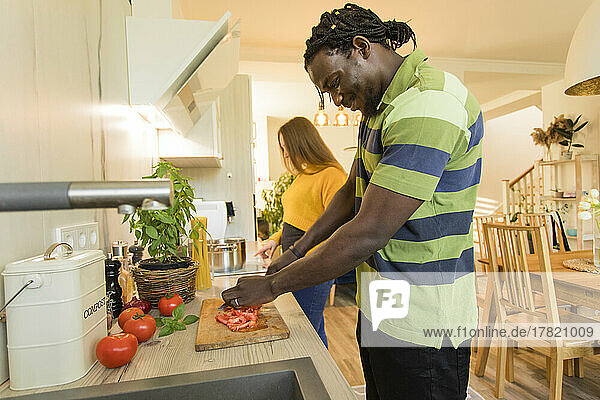 Smiling man cutting tomatoes by girlfriend in kitchen at home