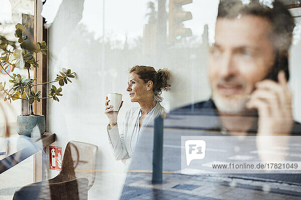 Businessman talking on mobile phone with businesswoman holding coffee cup in background seen through glass