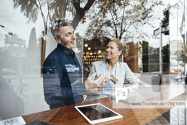 Smiling businesswoman holding credit card talking with businessman in cafe seen through window