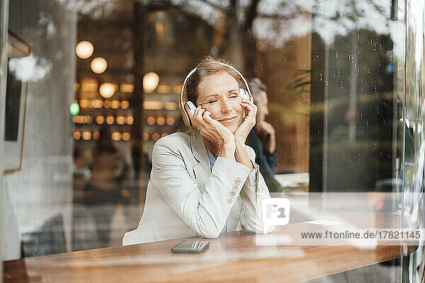 Smiling businesswoman with eyes closed listening music in cafe seen through glass