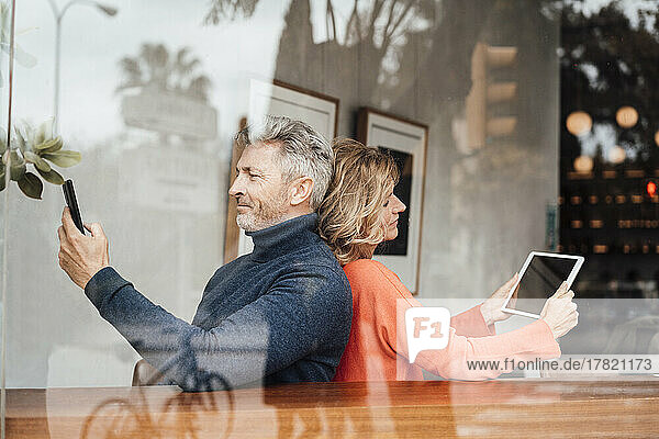 Smiling couple using wireless technologies sitting cafe seen through glass