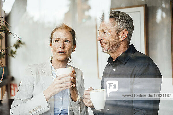 Businesswoman with businessman holding coffee cup in cafe seen through glass