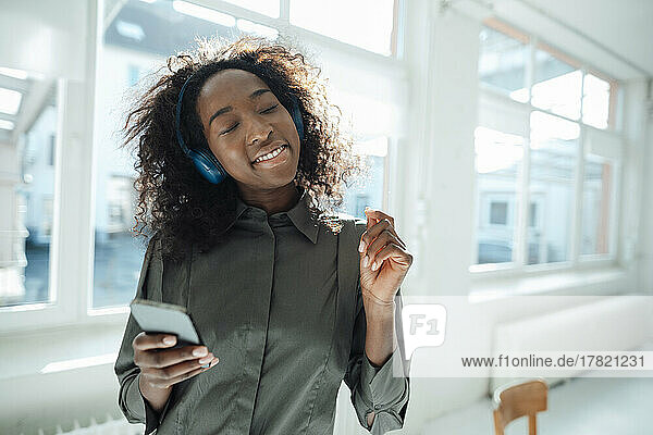 Young woman holding mobile phone enjoying music listening through wireless headphones in office