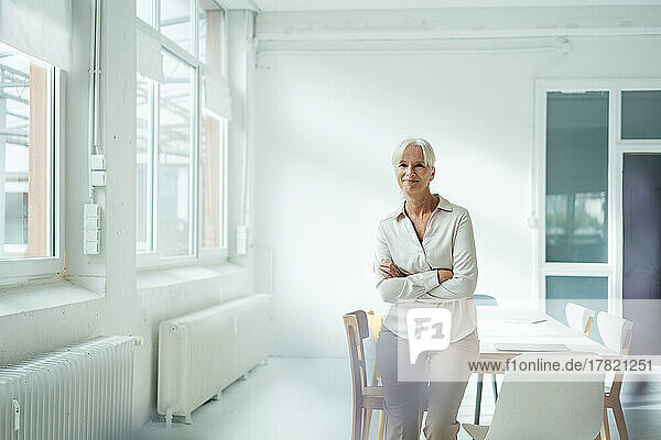 Smiling businesswoman with arms crossed leaning on desk in office