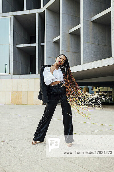 Selbstbewusste junge Geschäftsfrau mit langen geflochtenen braunen Haaren steht vor einem Bürogebäude