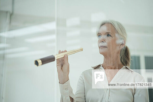 Senior businesswoman holding telescope in office seen through glass