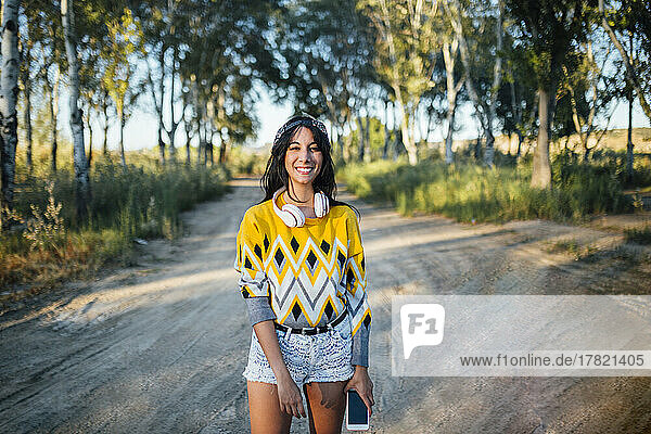 Smiling woman holding mobile phone on dirt road in nature