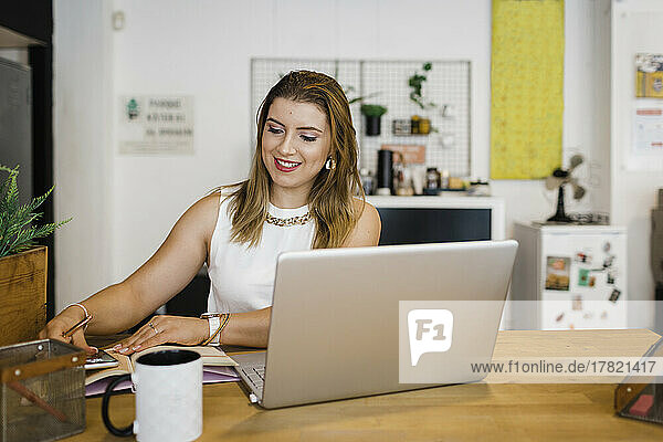 Smiling young businesswoman with smart phone and laptop sitting at desk in office