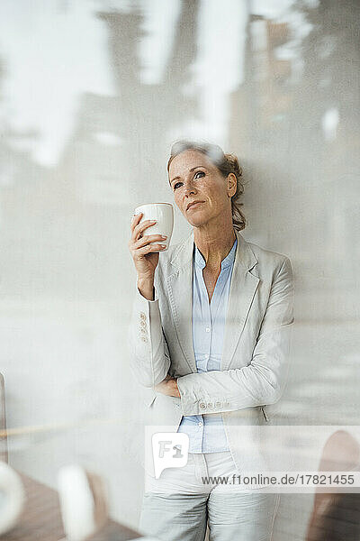 Thoughtful businesswoman holding coffee cup in cafe seen through glass