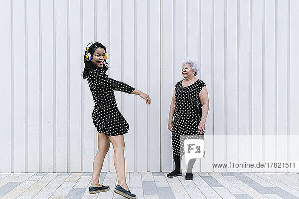 Senior woman looking at daughter listening music and dancing in front of white wall