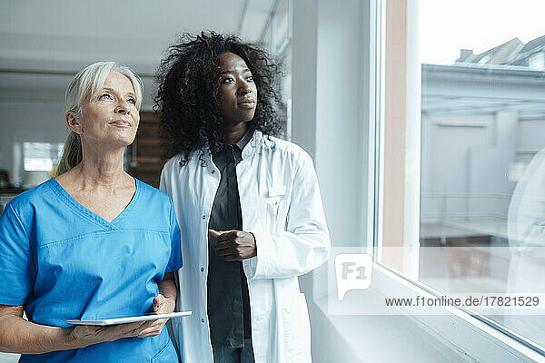 Krankenschwester hält Tablet-PC in der Hand und schaut neben Ärztin durchs Fenster