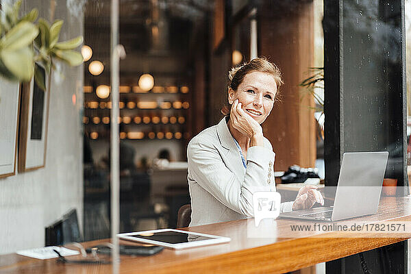 Smiling businesswoman sitting with head in hand at cafe seen through glass