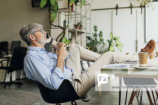 Mature businessman with disposable cup talking on smart phone in office