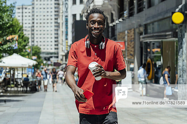 Happy man holding coffee cup and mobile phone walking in city on sunny day