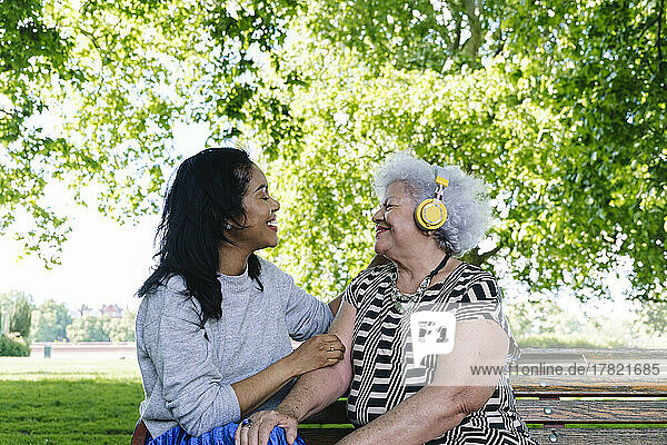 Mutter trägt kabellose Kopfhörer und sitzt mit Tochter auf Bank im Park
