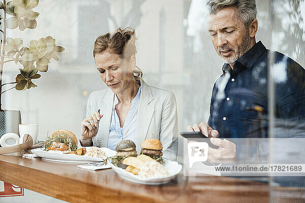 Businesswoman having lunch sitting by businessman using smart phone in cafe seen through glass