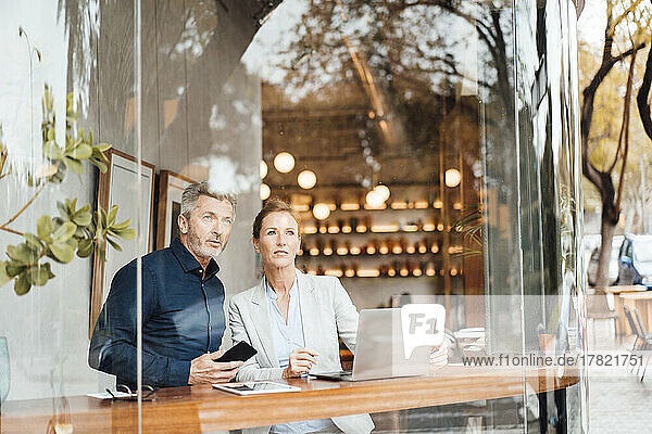Businessman holding mobile phone standing by businesswoman in cafe seen through window
