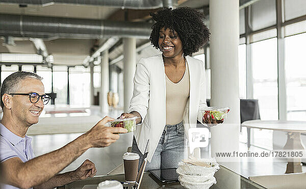 Happy young businesswoman giving salad box to businessman sitting at desk