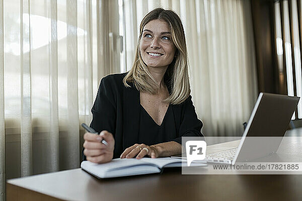 Smiling businesswoman holding pen sitting with laptop on table