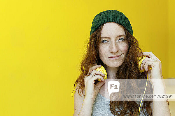 Woman in green knit hat holding headphones in front of yellow wall