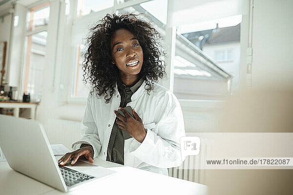 Female doctor holding mobile phone sitting with laptop at desk