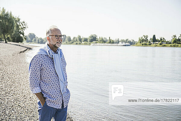 Senior man standing with hands in pockets at riverbank