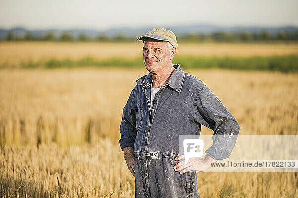 Smiling senior farmer wearing cap standing with arms akimbo at farm