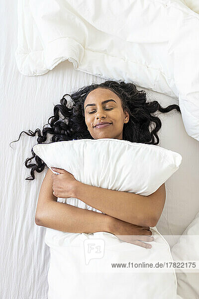 Happy woman with eyes closed embracing cushion on bed in bedroom