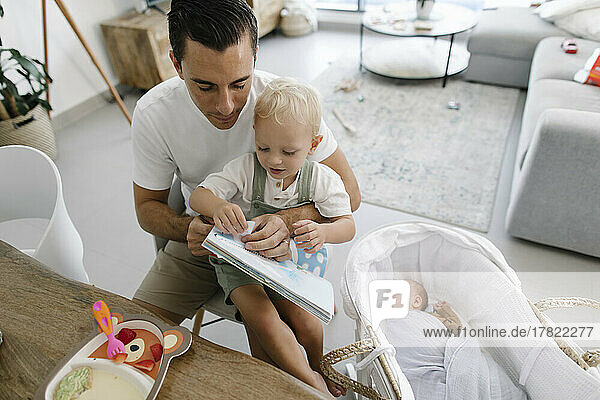 Father holding book sitting with son by baby sleeping in crib at home