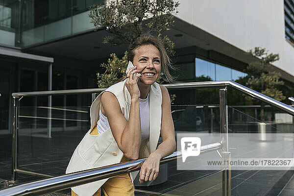 Smiling businesswoman talking through smart phone leaning on railing
