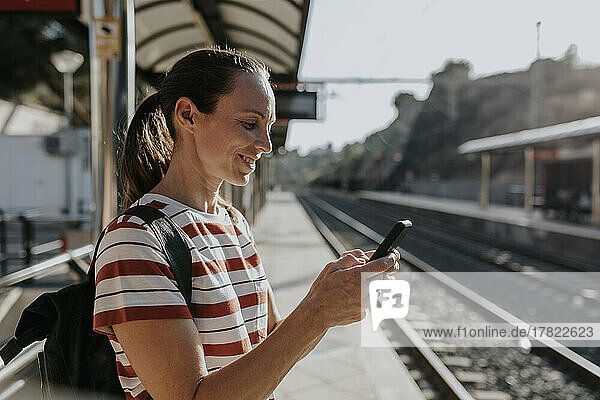 Smiling woman using smart phone standing at railroad station