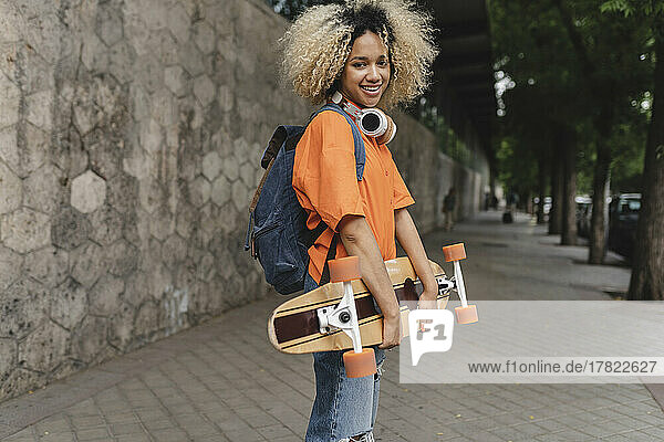 Smiling woman with backpack holding skateboard on footpath