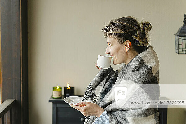 Smiling woman wrapped in blanket drinking coffee at home