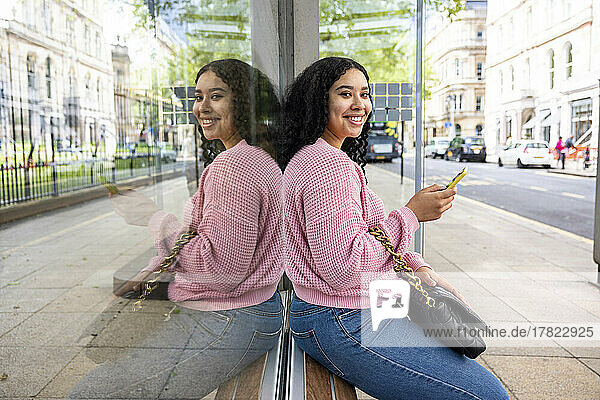 Happy young woman holding smart phone sitting on glass window