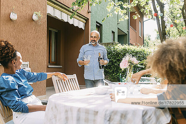 Smiling man holding wine bottle looking at woman sitting at dining table in backyard