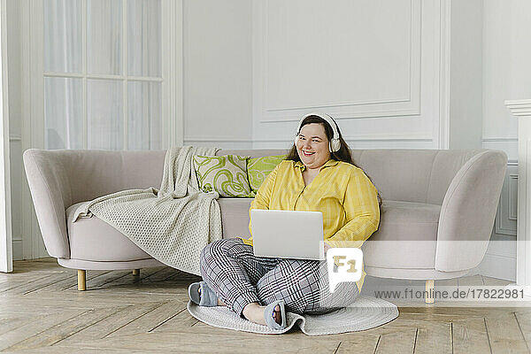 Smiling woman sitting with laptop enjoying music through wireless headphones at home
