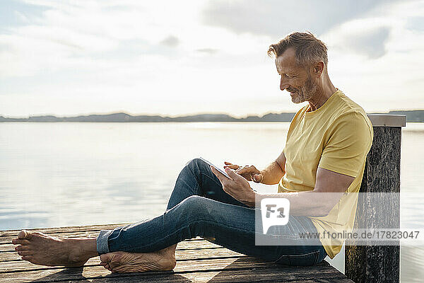 Mature man using tablet PC sitting on pier at lake