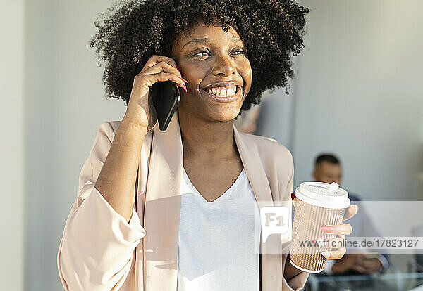 Happy businesswoman talking on smart phone holding disposable cup