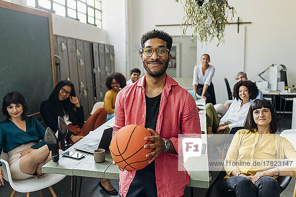 Smiling businessman with basketball by multiracial colleagues in office