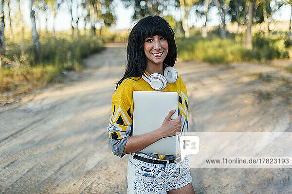 Smiling woman with bangs holding laptop standing on dirt road