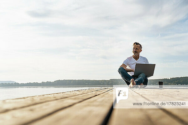 Thoughtful freelancer sitting with laptop on pier over lake