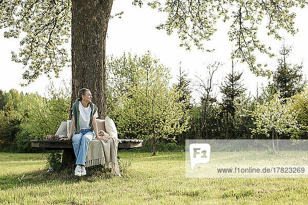 Smiling woman sitting on bench around tree trunk in garden