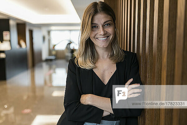 Smiling businesswoman with arms crossed standing by wooden wall at hotel