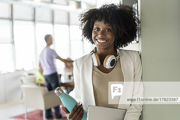 Smiling businesswoman with headphones holding water bottle at office