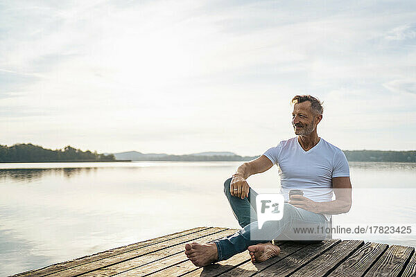 Lächelnder reifer Mann sitzt mit Einwegkaffeetasse auf dem Steg über dem See