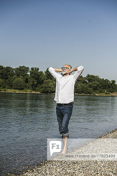 Älterer Mann steht mit den Händen hinter dem Kopf am Flussufer