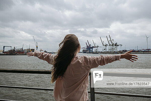 Young woman with arms outstretched standing by railing in front of Hamburg port